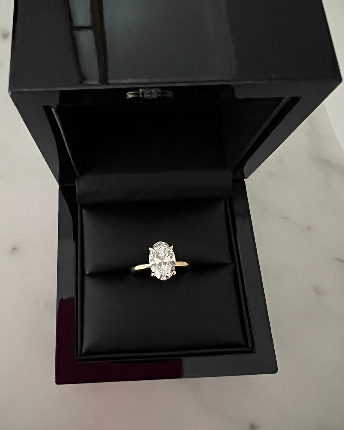 Oval diamond solitaire in yellow gold standing in its open black lacquer presentation box on marble