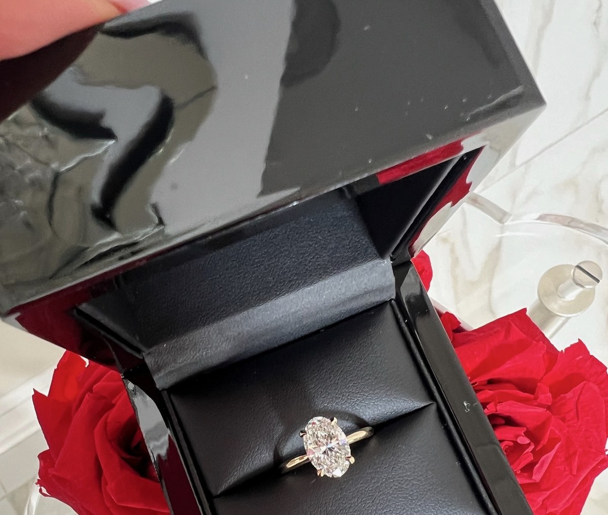Open black presentation box holding an oval diamond solitaire, held above a bouquet of red roses