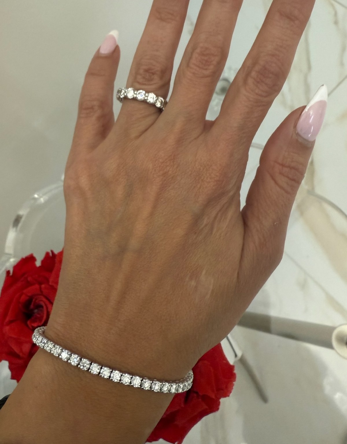 Round diamond tennis bracelet worn at the wrist beside a diamond band, photographed against marble and red roses